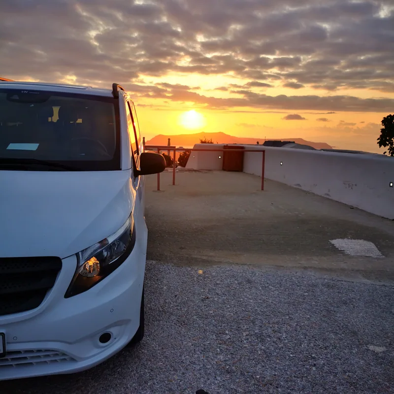 The VH Transfer van parked at a Santorini viewpoint as the sun sets over the caldera